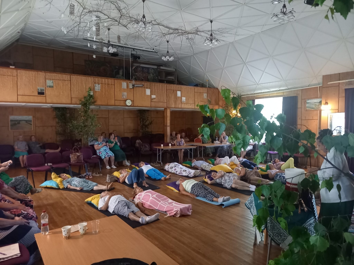 Seniori guļ uz paklājiņiem Tautas nama zālē, klausoties gonga mūziku