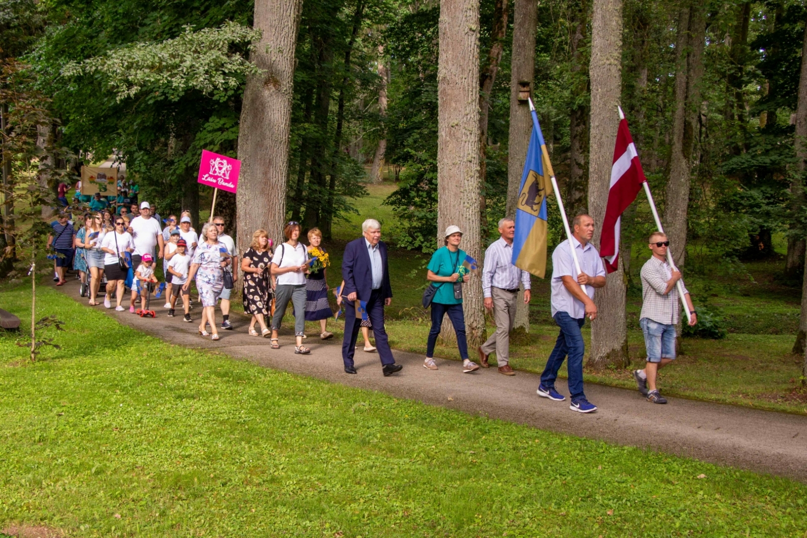 Cilvēki iet svētku gājienā, priekšā divi vīrieši ar Latvijas un Taurupes pagasta karogu Taurupes pagasta svētkos 07.2024.