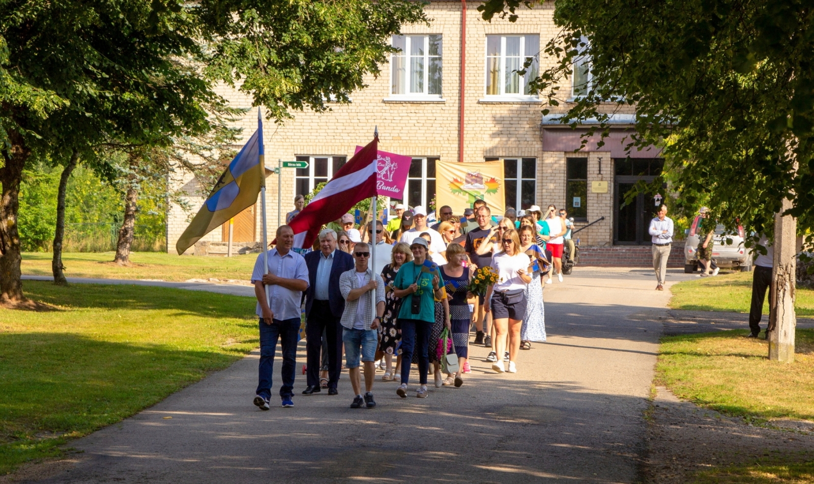 Cilvēki iet svētku gājienā, priekšā valsts karogs Taurupes pagasta svētkos 07.2024.