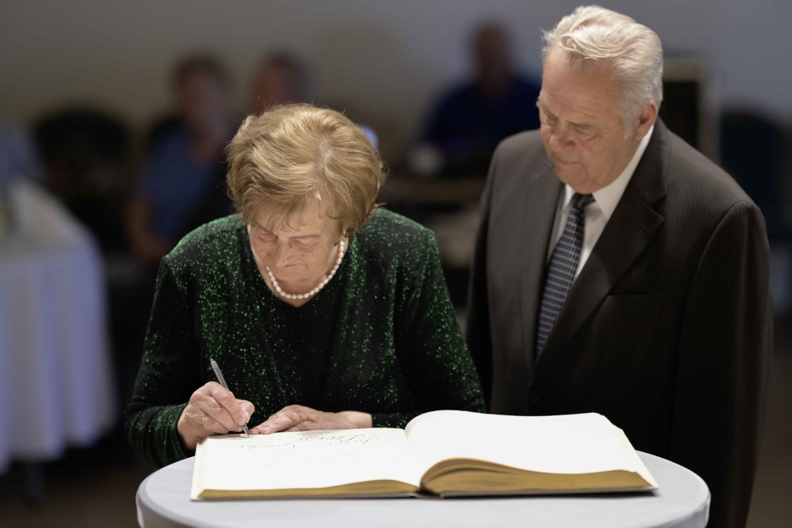 Sieviete zaļā tērpā parakstās Goda grāmatā, blakus vīrietis sirmiem matiem uzvalkā  Ogres novada Zelta kāzu pāris 31.05.2024